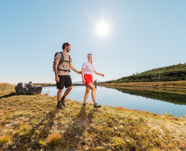 Muž a žena se drží za ruce na túře u jezera v Salcburském Lungau pod jasnou modrou oblohou.