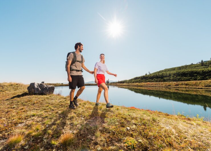 ©Salzburger Lungau (4) Muž a žena se drží za ruce na túře u jezera v Salcburském Lungau pod jasnou modrou oblohou.