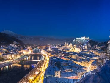 Zasněžené Město Salcburk v noci, osvětlené Wasserspiegelem, s Hohensalzburgem na Monchsbergu pod tmavě modrou oblohou.