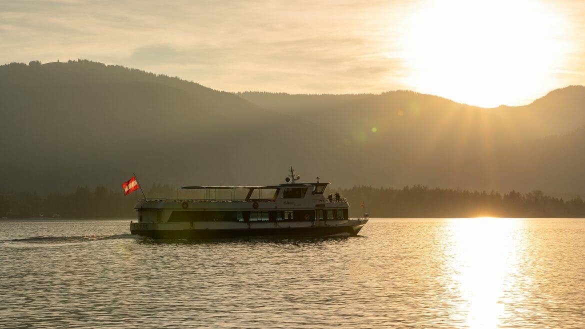 Za horami zapadá slunce, které se odráží ve vodní hladině jezera Wolfgangsee na němž pluje výletní loď