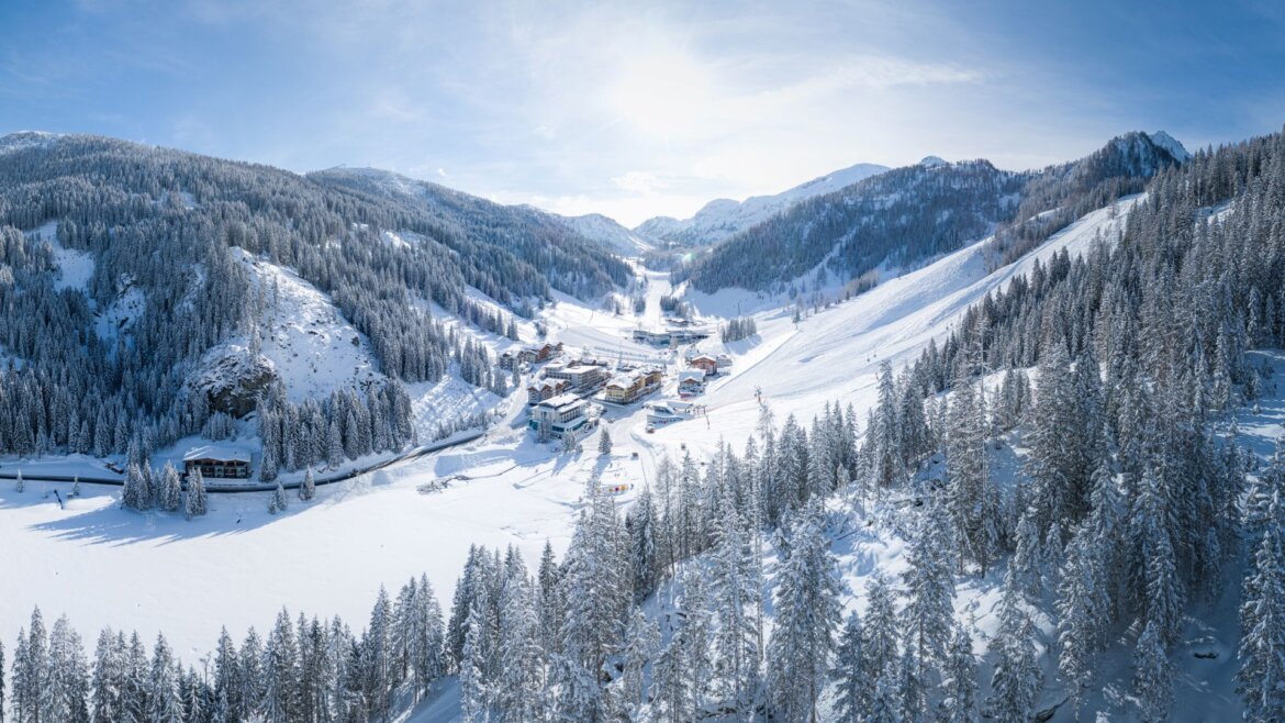 Zasněžená horská vesnička Altenmarkt-Zauchensee v zimě s borovými lesy a lyžařskými sjezdovkami pod modrou oblohou. (Zvětšený pohled)