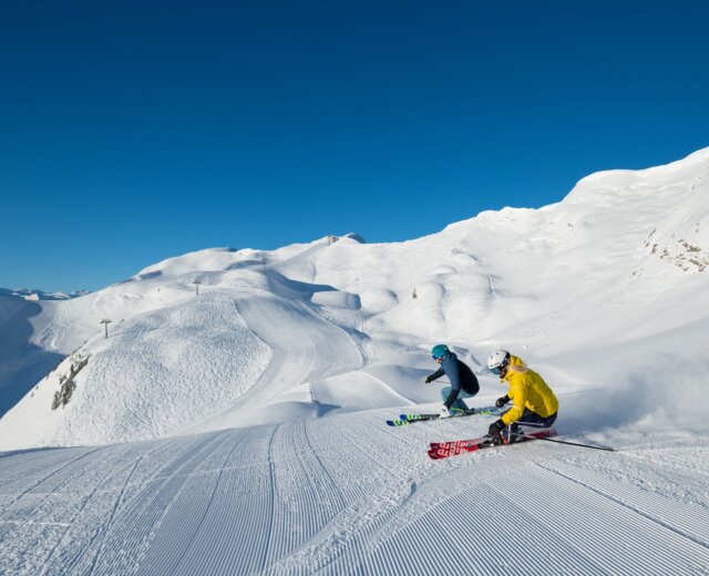 Raurisertal Rauriser Hochalmbahn Gipfel Blick auf Kolm Saigurn (c) TVB Rauris Fotograf Michael Gruber (2) Dva lyžaři v zářivém oblečení sjíždějí pod jasně modrou oblohou po upraveném zimním svahu v Raurisertalu.