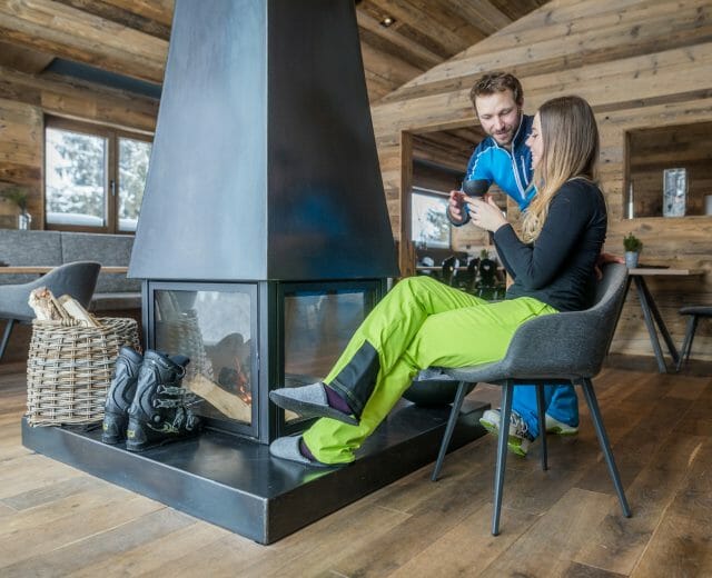 Pár v lyžařském oblečení odpočívá u moderního krbu v útulné dřevěné chatě, usmívá se a povídá si.