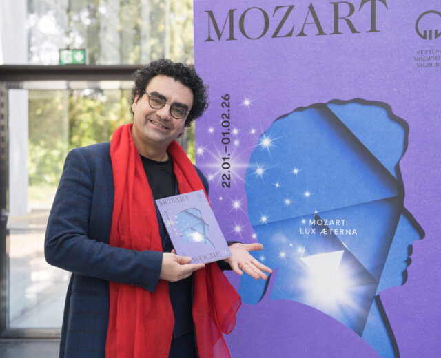 Rolando Villazon vedle plakátu události s modrou siluetou Mozarta