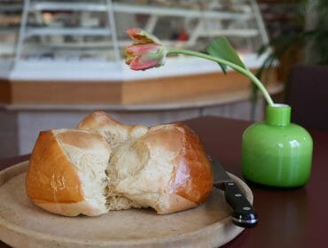 Kulatý bochník chleba na dřevěném prkénku s nožem, vedle růžové květiny v zelené váze.