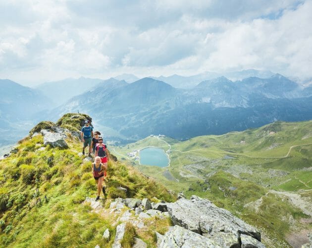 Turisté prochází po hřebeni hory v Obertauern v kulisách alpských štítů a malebného horského jezera