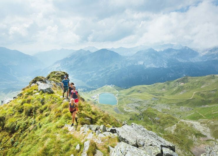 Turisté prochází po hřebeni hory v Obertauern v kulisách alpských štítů a malebného horského jezera