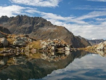 Turisté stojí na skalnatém terénu u průzračného horského jezera, v jehož hladině se odrážejí vrcholy a mraky.