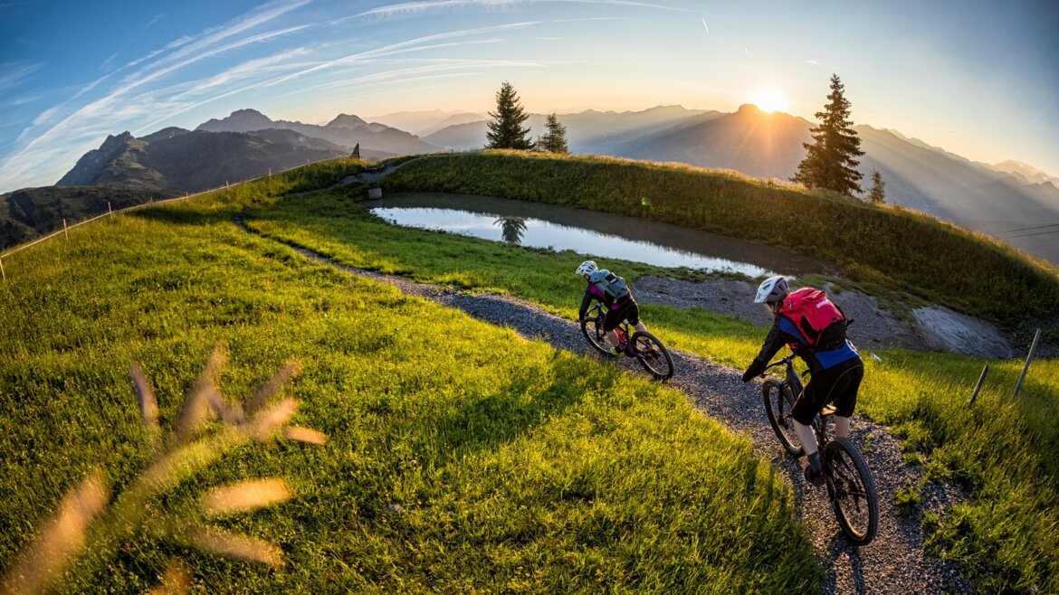 Dva cyklisté jedou při východu slunce z kopce v Grossarltal v Salcbursku v létě, rámované zelenou trávou a horami.