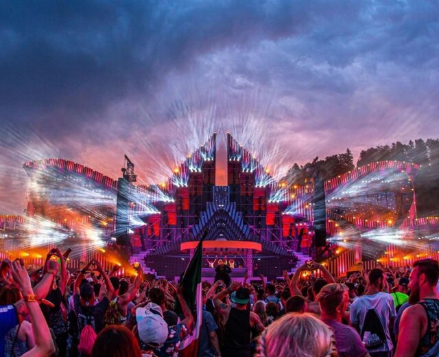 Dav na venkovním hudebním festivalu s barevnými světly na velkém pódiu a dramatickými mraky při západu slunce.