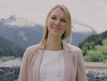 Žena se usmívá venku se zasněženými horami a zelenými kopci v pozadí v zamračeném dni.