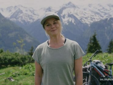 Žena v čepici se usmívá venku s horami a kolem v pozadí.