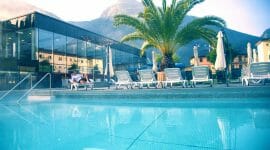 _MAX9519 Venkovní bazén s lehátky, palmou, prosklenou budovou a horskou kulisou za slunečného dne.