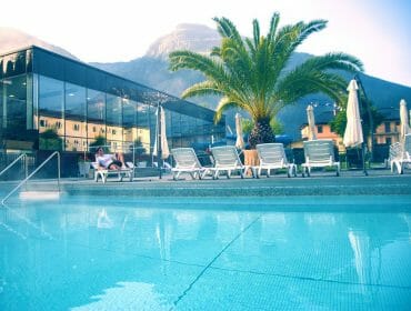 Venkovní bazén s lehátky, palmou, prosklenou budovou a horskou kulisou za slunečného dne.