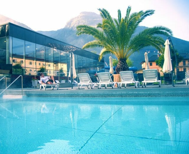 Venkovní bazén s lehátky, palmou, prosklenou budovou a horskou kulisou za slunečného dne.