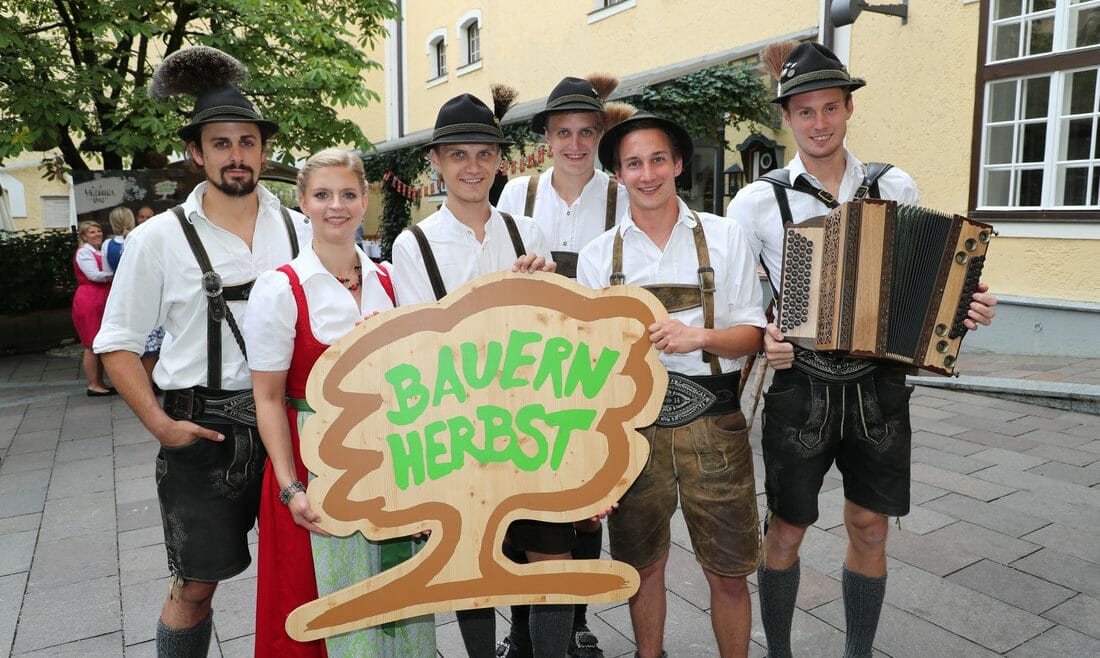 Účastníci v krojích drží logo Farmářského podzimu, symbol stromu se zeleným nápisem Bauernherbst