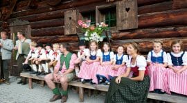 Bauernherbst – Kinder auf Bank vor Hütte Farmářský podzim a dožínky v Salcbursku jsou plné tradičních akcí. Děti v krojích sedí na lavičce před dřevěnou chalupou a poslouchají lidovou hudbu.