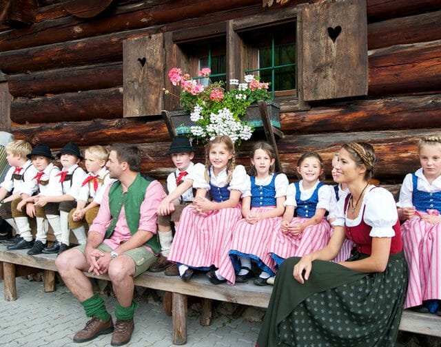 Bauernherbst – Kinder auf Bank vor Hütte Farmářský podzim a dožínky v Salcbursku jsou plné tradičních akcí. Děti v krojích sedí na lavičce před dřevěnou chalupou a poslouchají lidovou hudbu.