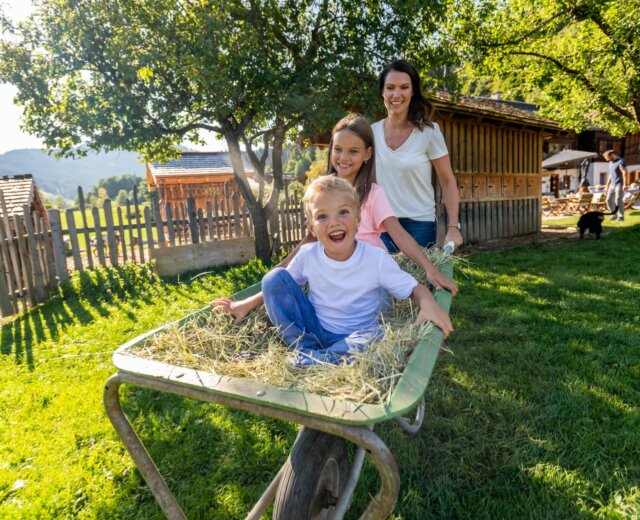 Žena veze smějící se děti zahradou na kolečku se slámou.