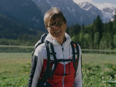 Video thumbnail for youtube video ykqrqdczfbq Usměvavá starší žena v turistickém oblečení stojí na zeleném poli s horami a stromy v pozadí.