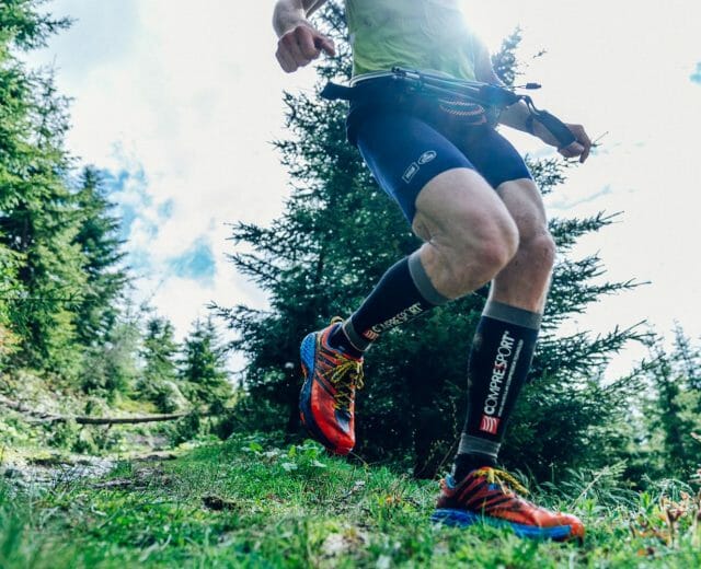 Běžec v barevných botách a sportovním oblečení běží po lesní cestě obklopené zelenými stromy a trávou.