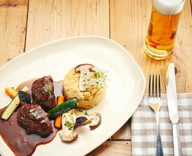vc_0421_website_bilder_28 Talíř s hovězím masem v omáčce, zeleninou, polentou s houbami a pivem na stole Via Culinaria hostince.