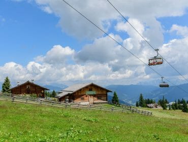 Dvě dřevěné chaty na travnatém svahu v obci Ski amadé dovolená, s lyžařským vlekem a horami pod modrou oblohou.