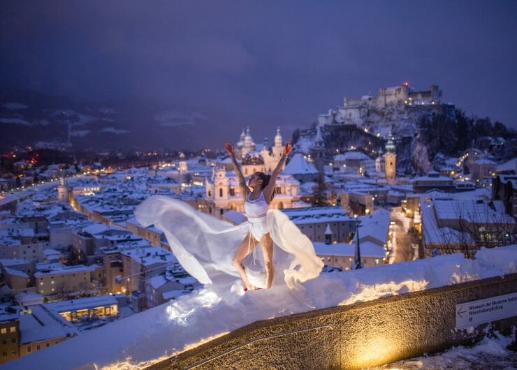 Tanečnice s vlající bílou sukní na promenádě nad nočním, historickým centrem Salcburku s jeho zasněženými střechami a osvětlenými památkami