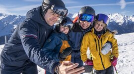 Rodina v lyžařské výstroji se usmívá a pořizuje si selfie na Zimní rodinná dovolená Wildkogel Arena s modrou oblohou.