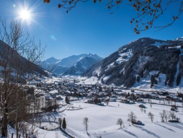 Raurisertal_Talansicht_Lila&Plietsch Zasněžená vesnice Raurisertal, zasazená v údolí pod jasným sluncem.