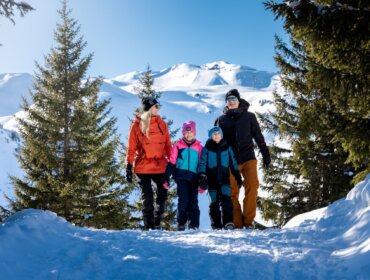 Raurisertal_Winterwanderweg Hochalm ©Hochalmbahnen AG_final (9) Čtyřčlenná rodina kráčí po zasněžené stezce v Raurisertalu, obklopená stromy a horami.
