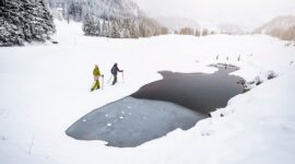 SLT_Regionsseite_OT_CZ_Abseits der Piste Dva lidé kráčí s lyžařskými hůlkami poblíž zamrzlého rybníka v Obertauernu mimo sjezdovky, v pozadí jsou zasněžené vrcholky hor.