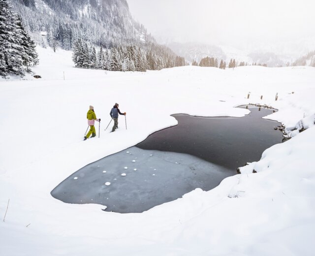 SLT_Regionsseite_OT_CZ_Abseits der Piste Dva lidé kráčí s lyžařskými hůlkami poblíž zamrzlého rybníka v Obertauernu mimo sjezdovky, v pozadí jsou zasněžené vrcholky hor.