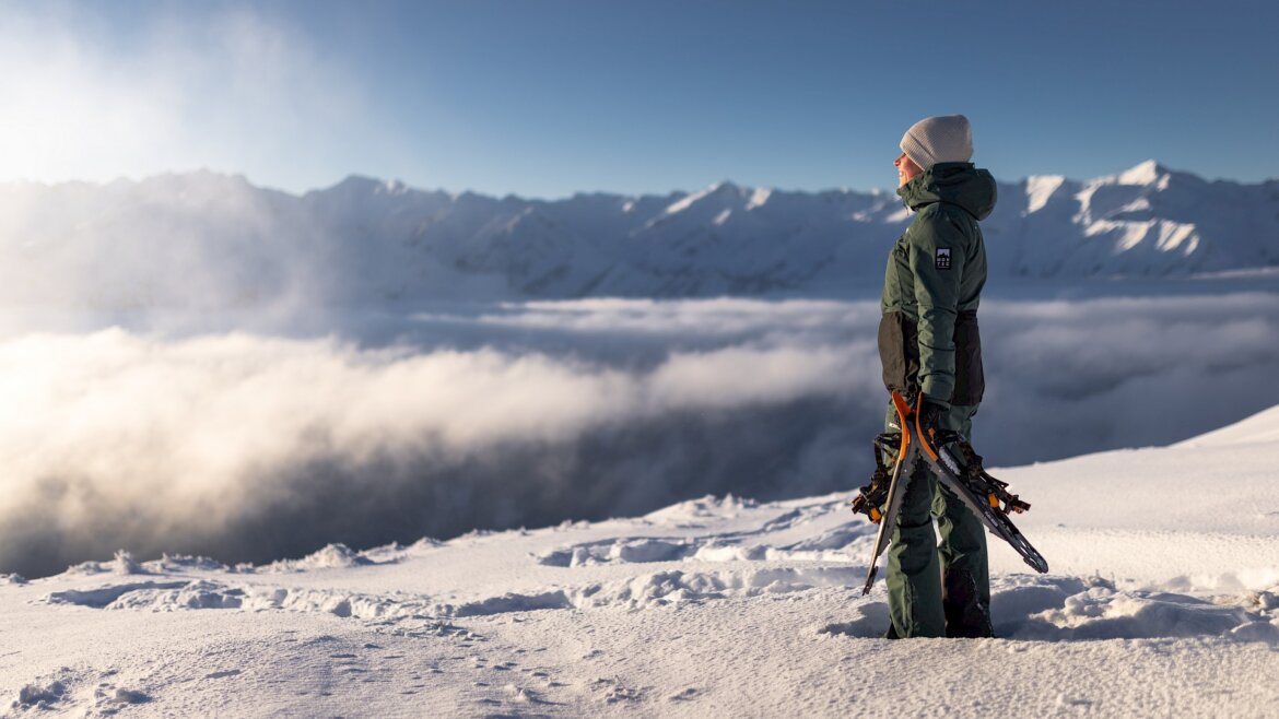 Osoba v zimní výstroji stojí na zasněžené hoře, připravená na zimní turistiku Wildkogel Arena při východu slunce.