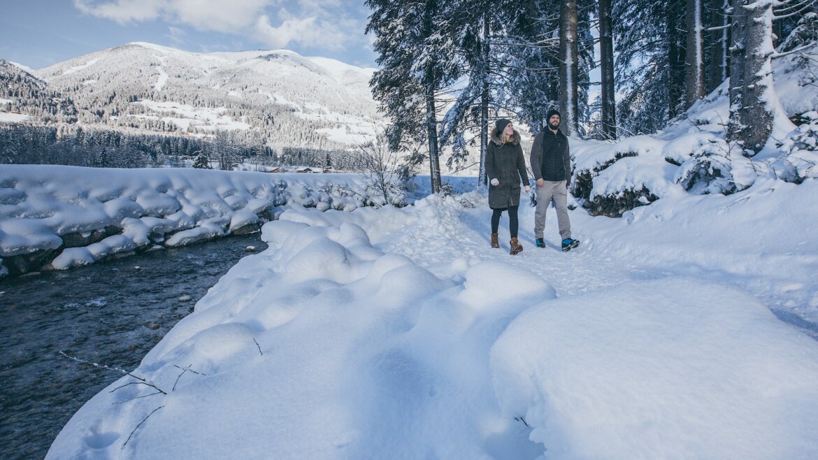 Dva lidé se procházejí po zimní turistické stezce u zasněžené řeky v zimní krajině Wildkogel Areny.