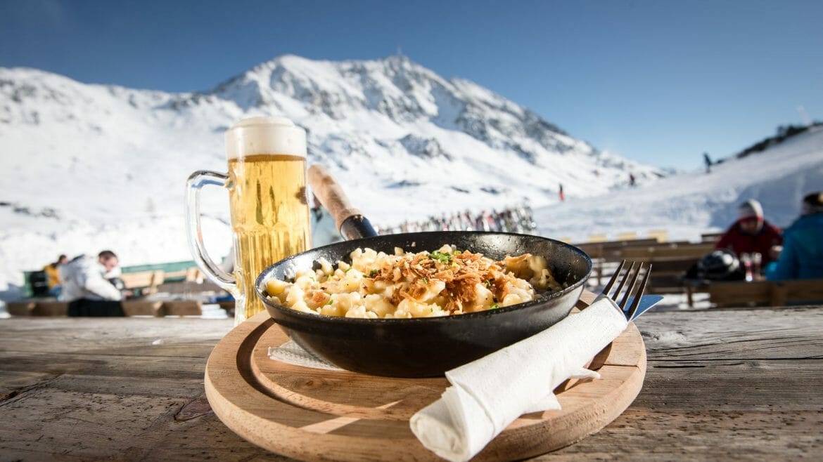 Půllitr točeného piva, pánvička plná noků se sýrovou omáčkou a smaženou cibulkou, to vše korunované výhledem na zasněžené vrcholy hor v Obertauern