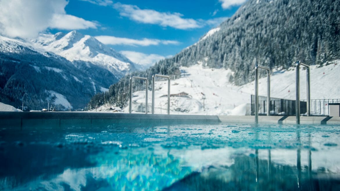 Felsentherme Bad Gastein im Winter Venkovní bazén s průzračně modrou vodou, obklopený zasněženými horami pod jasnou, částečně zataženou oblohou.