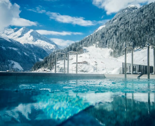 Felsentherme Bad Gastein im Winter Venkovní bazén s průzračně modrou vodou, obklopený zasněženými horami pod jasnou, částečně zataženou oblohou.
