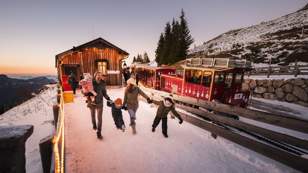 Rodina se prochází po zasněžené zemi v blízkosti červeného horského vláčku, dřevěné chaty a Turistické cíle Salcburk a Solná komora.