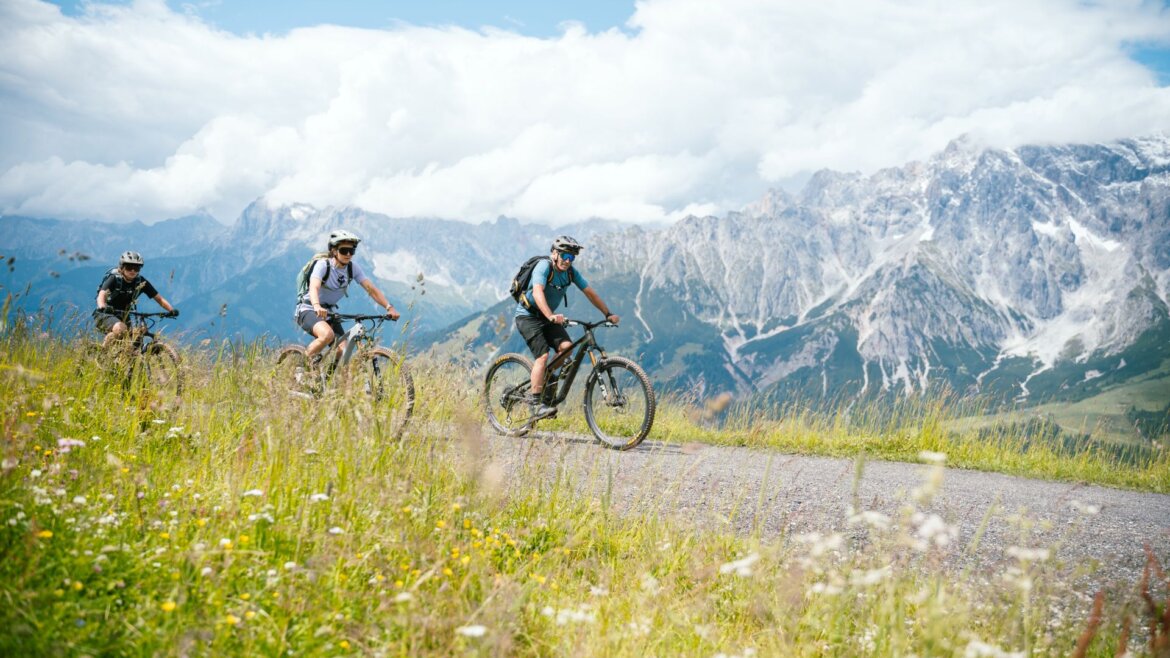 Tři cyklisté jedou na MTB kolech po stezce přes travnaté kopce s horami v oblasti Hochkönig horská kola. (Zvětšený pohled)