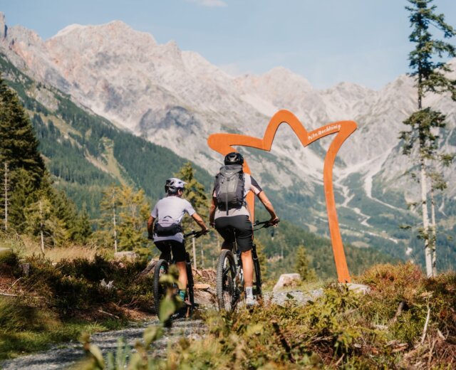 Biken_Primbach_Hochkönig Dva cyklisté jedou po flowtrailu na sjezdovce Hochkönig směrem k velkému oranžovému rámu s malebnými vrcholy za zády.