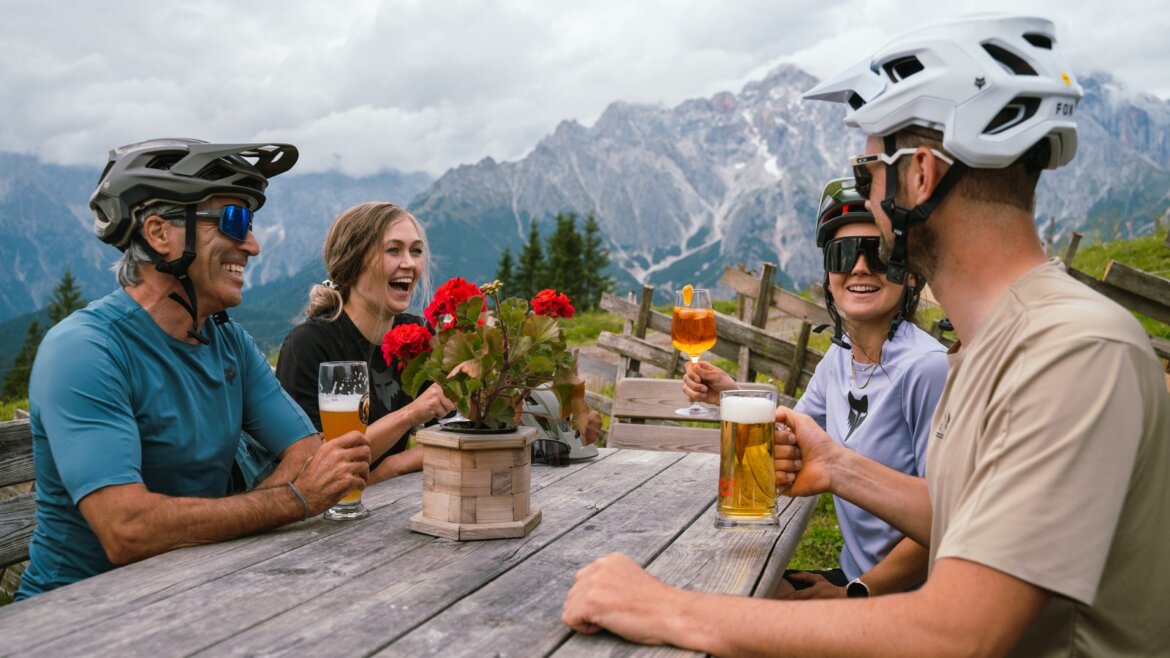 Čtyři cyklisté na MTB v helmách odpočívají u piknikového stolu a smějí se s horami Hochkönig za zády.