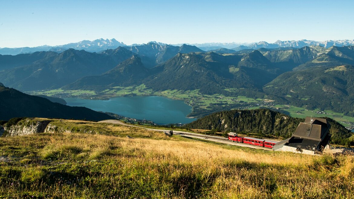 Zubačka Schafbergbahn stoupá po horském svahu s výhledem na jezero a vzdálené vrcholy pod modrou oblohou. (Zvětšený pohled)