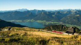 Zubačka Schafbergbahn stoupá po horském svahu s výhledem na jezero a vzdálené vrcholy pod modrou oblohou.