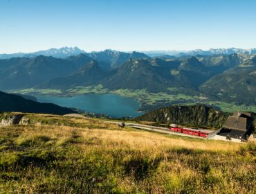 marco_riebler-3994 Zubačka Schafbergbahn stoupá po horském svahu s výhledem na jezero a vzdálené vrcholy pod modrou oblohou.