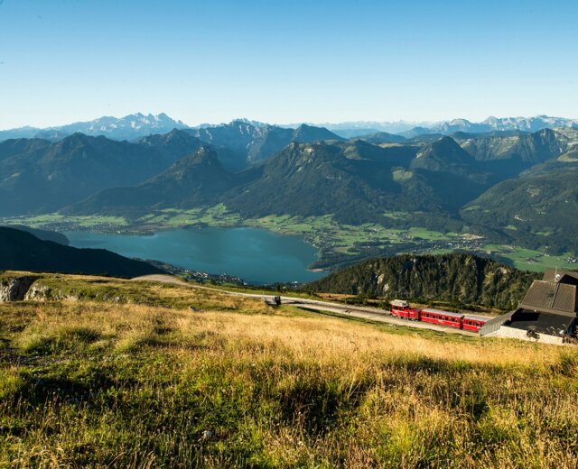 Zubačka Schafbergbahn stoupá po horském svahu s výhledem na jezero a vzdálené vrcholy pod modrou oblohou.