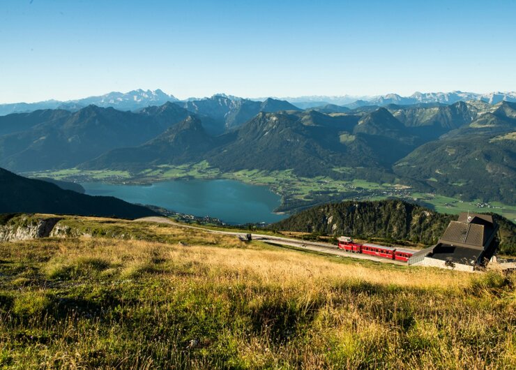 Zubačka Schafbergbahn stoupá po horském svahu s výhledem na jezero a vzdálené vrcholy pod modrou oblohou.