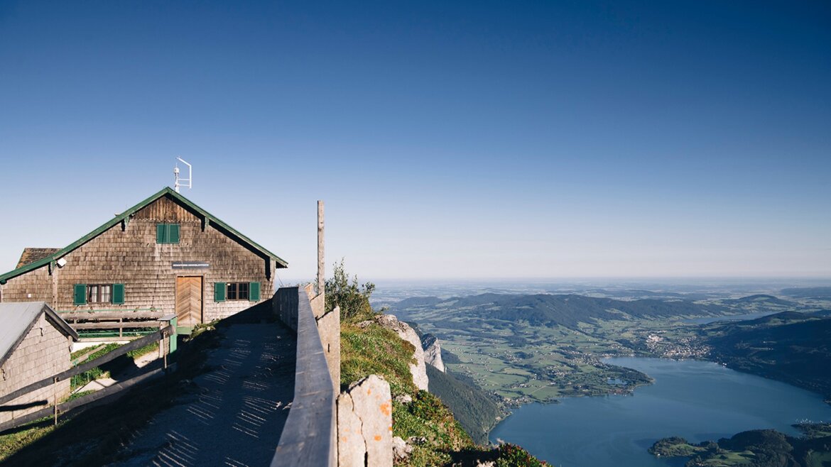 Okouzlující pohled na krajinu a Mondsee od Himmelspforte ze Schafbergu © Salzburg AG Tourismus GmbH Dřevěná chata Himmelspforte na Schafbergu s výhledem na jezero Mondsee a údolí pod jasnou modrou oblohou. (Zvětšený pohled)