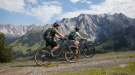 Dva cyklisté jedou na elektrokole Hochkönig po štěrkové cestě s malebnými horami a zelenými údolími v pozadí.