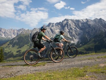 medium-Paerchen-geniesst-Tour-mit-dem-eBike-vor-herrlichem-Bergpanorama Dva cyklisté jedou na elektrokole Hochkönig po štěrkové cestě s malebnými horami a zelenými údolími v pozadí.